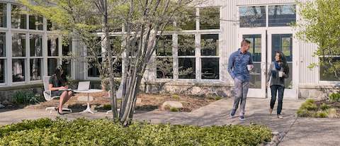 A young woman and man walk through a courtyard outside, chatting and drinking coffee while a woman works on a laptop nearby. A young woman and man walk through a courtyard outside, chatting and drinking coffee while a woman works on a laptop nearby.