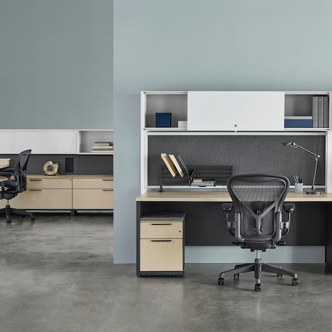 A Canvas Metal Desk with white upper storage, gray fabric back panels, and light wood surfaces with another workstation in the background. A Canvas Metal Desk with white upper storage, gray fabric back panels, and light wood surfaces with another workstation in the background.