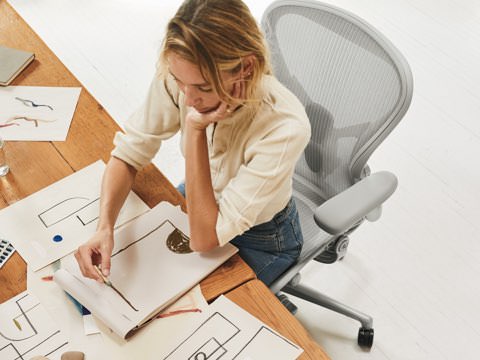 A woman sitting at a desk in her Aeron Chair, sketching shapes and designs on paper. A woman sitting at a desk in her Aeron Chair, sketching shapes and designs on paper.