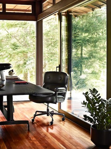 A black leather Eames Executive Chair with a black AGL table in a glass-walled home office overlooking trees. A black leather Eames Executive Chair with a black AGL table in a glass-walled home office overlooking trees.