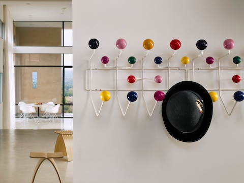 A man's hat hangs from one of two adjacent Eames Hang-It-All storage racks, both with a white wire frame and multicolored knobs. A man's hat hangs from one of two adjacent Eames Hang-It-All storage racks, both with a white wire frame and multicolored knobs.
