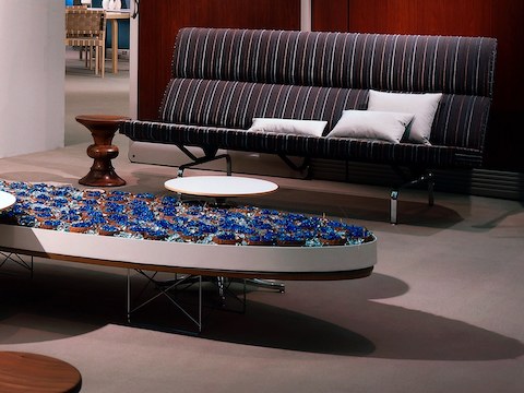 An Eames Sofa Compact with a striped fabric next to an Eames Walnut Stool in a workplace lounge. An Eames Sofa Compact with a striped fabric next to an Eames Walnut Stool in a workplace lounge.
