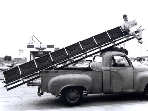 Vintage photo of a man sitting at the end of an Eames Tandem Sling Seating run being delivered via pickup truck. Vintage photo of a man sitting at the end of an Eames Tandem Sling Seating run being delivered via pickup truck.