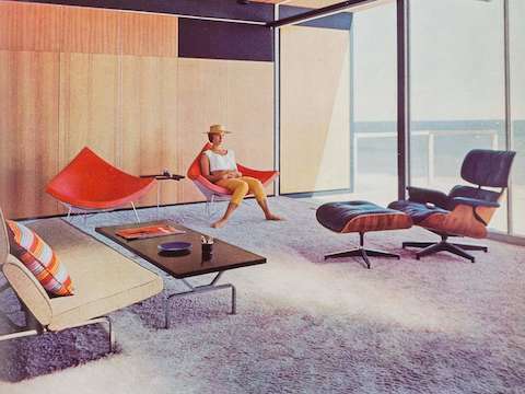 A woman sits in one of a pair of orange-pink Nelson Coconut Chairs in the sunny living room of a mid-century modern home in Malibu. An Eames Lounge Chair and Ottoman and Eames Sofa Compact are nearby. A woman sits in one of a pair of orange-pink Nelson Coconut Chairs in the sunny living room of a mid-century modern home in Malibu. An Eames Lounge Chair and Ottoman and Eames Sofa Compact are nearby.