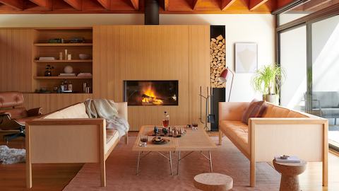 A living space featuring the Nelson Cube Sofa, Nelson Cane Bench, and Eames Lounge Chair accessorized with cork stools, a floor lamp, linen rug, chess set, and fireplace tools. A living space featuring the Nelson Cube Sofa, Nelson Cane Bench, and Eames Lounge Chair accessorized with cork stools, a floor lamp, linen rug, chess set, and fireplace tools.