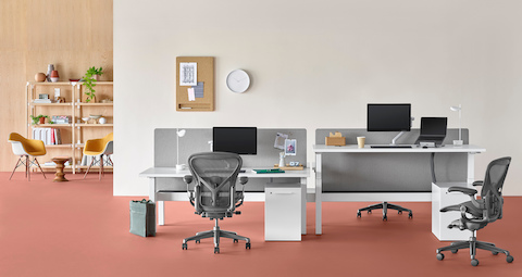 An office setting with a Nevi Link standing desk system in the foreground with white rectangular work surfaces and black Aeron office chairs, and a lounge area with Eames shell chairs and bookcase in the background. One of the four desks is raised to standing height. An office setting with a Nevi Link standing desk system in the foreground with white rectangular work surfaces and black Aeron office chairs, and a lounge area with Eames shell chairs and bookcase in the background. One of the four desks is raised to standing height.