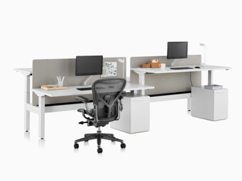 A Nevi Link standing desk system with a black Aeron office chair, privacy screens, monitors, and rectangular work surfaces. Two of the four desks are raised to standing height. A Nevi Link standing desk system with a black Aeron office chair, privacy screens, monitors, and rectangular work surfaces. Two of the four desks are raised to standing height.