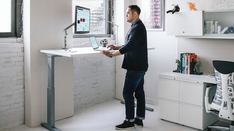 A man works on a computer while standing at a rectangular Renew Sit-to-Stand Table. A man works on a computer while standing at a rectangular Renew Sit-to-Stand Table.