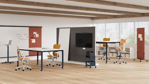 Open plan agile team space shown with two 4-leg height adjustable meeting tables featuring white surfaces, dark blue legs and technology buckets in center of tables. Room is split by sub-architectural boundary that features a monitor for video conferencing. Open plan agile team space shown with two 4-leg height adjustable meeting tables featuring white surfaces, dark blue legs and technology buckets in center of tables. Room is split by sub-architectural boundary that features a monitor for video conferencing.