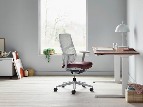 A Verus Chair with a dark red upholstered seat and white Triflex back next to a Renew Sit-to-Stand Table in a home office with gray walls and light hardwood floors. A Verus Chair with a dark red upholstered seat and white Triflex back next to a Renew Sit-to-Stand Table in a home office with gray walls and light hardwood floors.