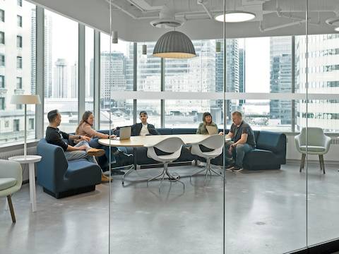 Five people sit on blue Chadwick Modular Seating in a glass-walled conference room. Two white Nelson Swag Leg chairs sit across the table from them, and a gray Saiba Chair sits off to the side. Five people sit on blue Chadwick Modular Seating in a glass-walled conference room. Two white Nelson Swag Leg chairs sit across the table from them, and a gray Saiba Chair sits off to the side.