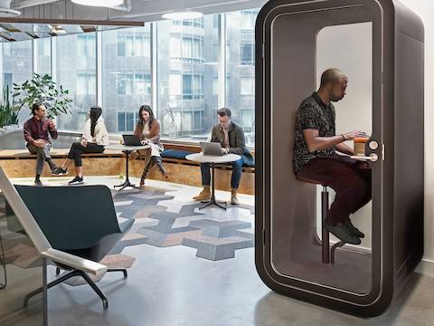 A person sits on a stool in a Framery O Office Phone Booth. Nearby, four people sit, chat, and work on laptops in a window seat. A person sits on a stool in a Framery O Office Phone Booth. Nearby, four people sit, chat, and work on laptops in a window seat.