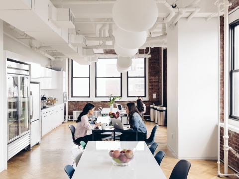 Two women sit and talk inside of an office cafe area full of natural light. Two women sit and talk inside of an office cafe area full of natural light.