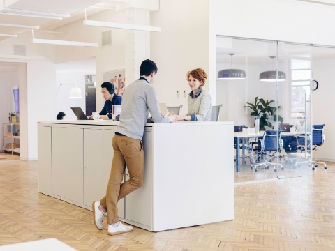Office employees work and talk at an extended desk setup. Office employees work and talk at an extended desk setup.