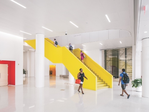 People inside of a library lobby, with a bold yellow staircase at the center. People inside of a library lobby, with a bold yellow staircase at the center.
