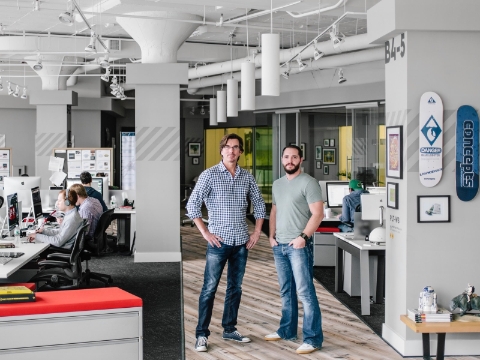 Two men pose in an office space while others work at their desks in the background. Two men pose in an office space while others work at their desks in the background.