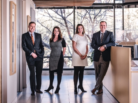 Four people pose in front of a window inside their office space. Four people pose in front of a window inside their office space.