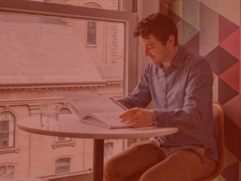 A person sits at a high top desk and reads a book. The surrounding office is bright and airy, and is outfitted with design elements that reflect the company's brand. A person sits at a high top desk and reads a book. The surrounding office is bright and airy, and is outfitted with design elements that reflect the company's brand.