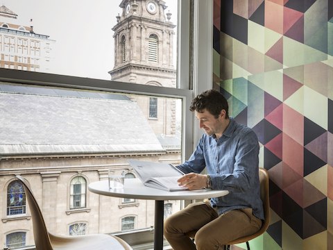 In a private setting overlooking a stone building, a man reviews a book while seated at a round table in an Eames Molded Wood Chair. In a private setting overlooking a stone building, a man reviews a book while seated at a round table in an Eames Molded Wood Chair.