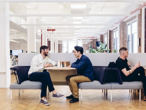 Co-workers interact in a casual space featuring a peninsula table and Public Office Landscape seating in shades of blue and gray. Co-workers interact in a casual space featuring a peninsula table and Public Office Landscape seating in shades of blue and gray.