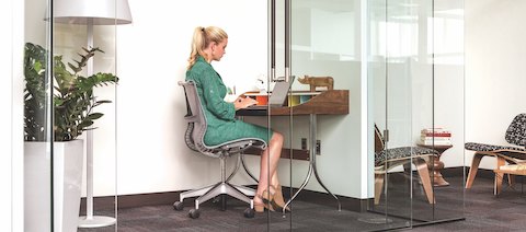 A person in green dress is sitting in a Setu Chair and typing on her laptop at a small desk in a glass-walled workspace. Spaces like this give people the opportunity to foucs on heads-down work in an open office setting. A person in green dress is sitting in a Setu Chair and typing on her laptop at a small desk in a glass-walled workspace. Spaces like this give people the opportunity to foucs on heads-down work in an open office setting.