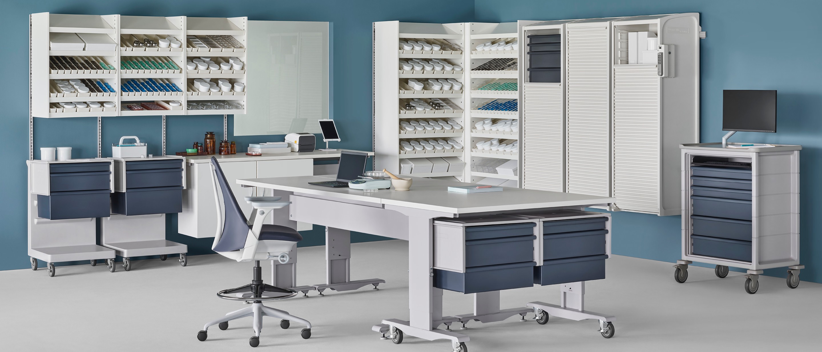 A pharmacy environment containing a Co/Struc System of dispensing shelves, lockers, process tables, L Carts with blue drawers, a technology cart with blue drawers, and a Sayl Stool. A pharmacy environment containing a Co/Struc System of dispensing shelves, lockers, process tables, L Carts with blue drawers, a technology cart with blue drawers, and a Sayl Stool.
