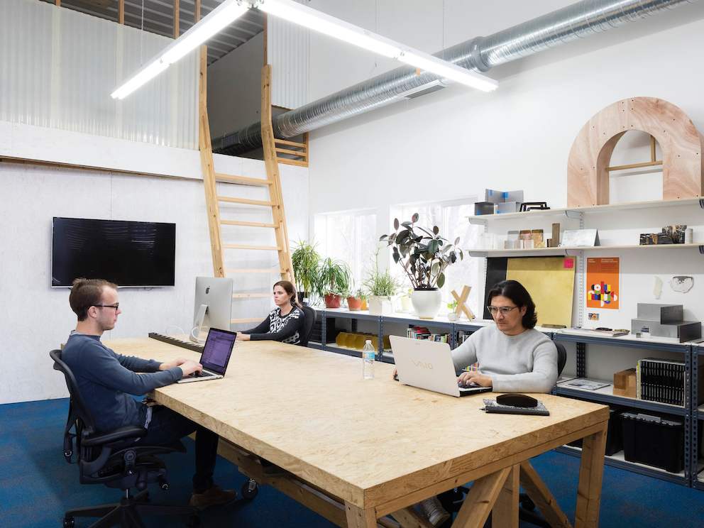 A woman and two men work on computers at a large table in a bright studio. A woman and two men work on computers at a large table in a bright studio.