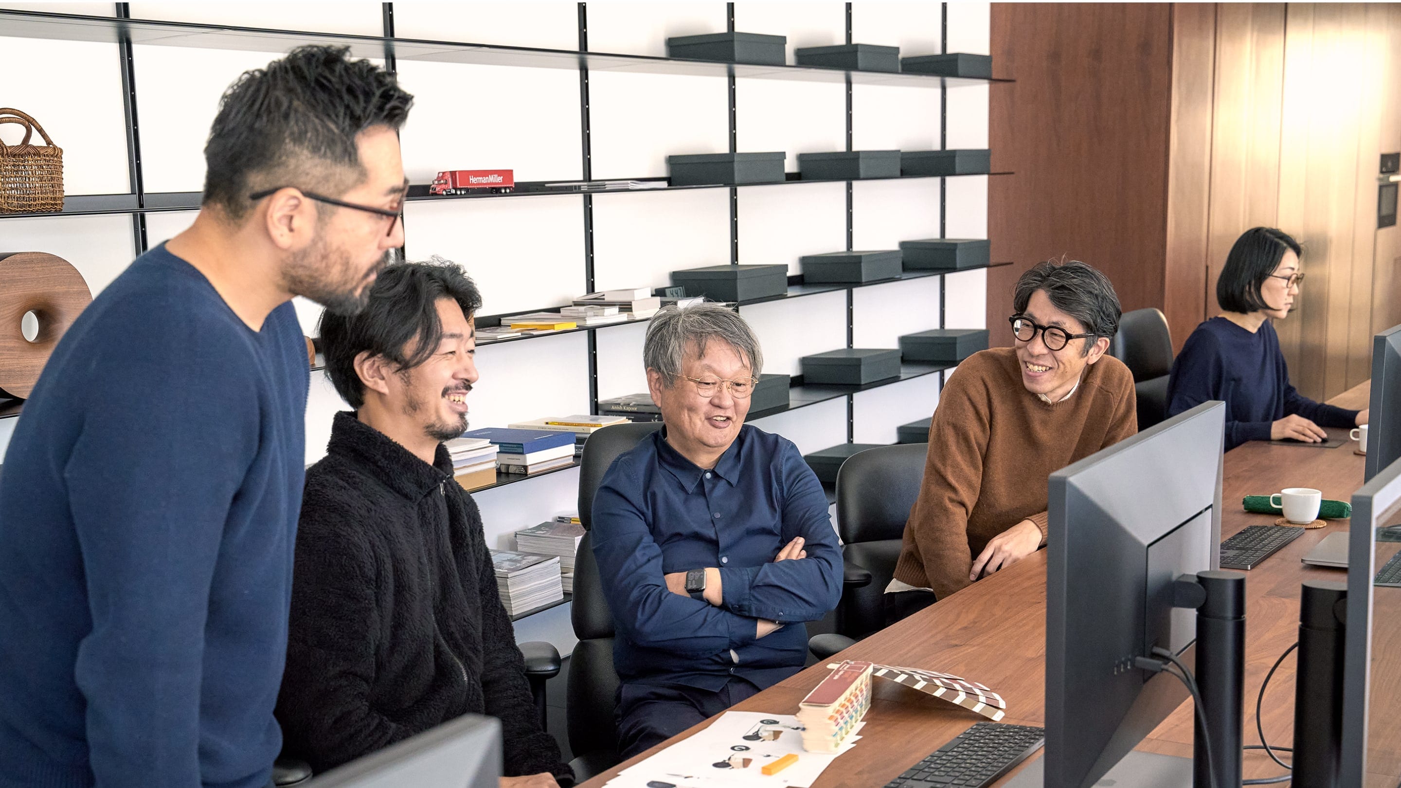 Naoto Fukasawa sits in the center of a group of five designers at a long wooden desk, with a minimal black shelf behind them featuring wooden sculptures, various ephemera, and black file boxes. In front of them are a Pantone swatch deck and computer monitors. Naoto Fukasawa sits in the center of a group of five designers at a long wooden desk, with a minimal black shelf behind them featuring wooden sculptures, various ephemera, and black file boxes. In front of them are a Pantone swatch deck and computer monitors.