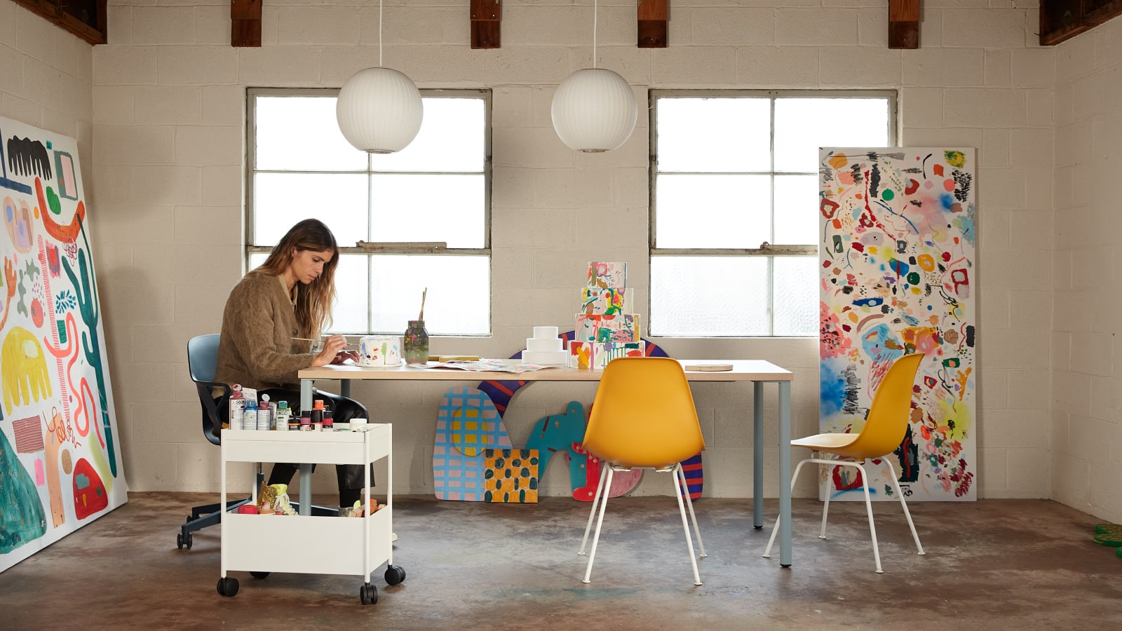 Artist Kindah Khalidy sitting in a Zeph Chair at a table, with two yellow Eames shell chairs. Artist Kindah Khalidy sitting in a Zeph Chair at a table, with two yellow Eames shell chairs.