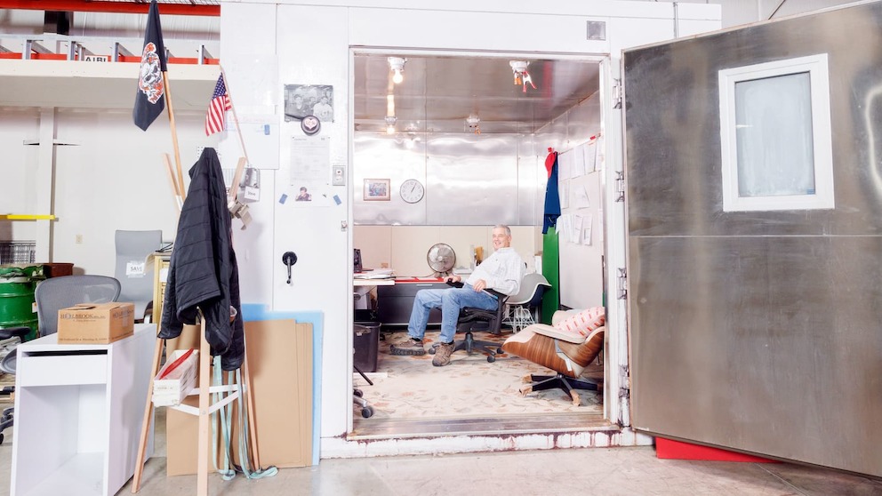 A large white chamber sits inside the Test Lab warehouse, with its aluminum door opened wide. Inside is a makeshift office space, where a man sits in his office chair at a desk. A large white chamber sits inside the Test Lab warehouse, with its aluminum door opened wide. Inside is a makeshift office space, where a man sits in his office chair at a desk.