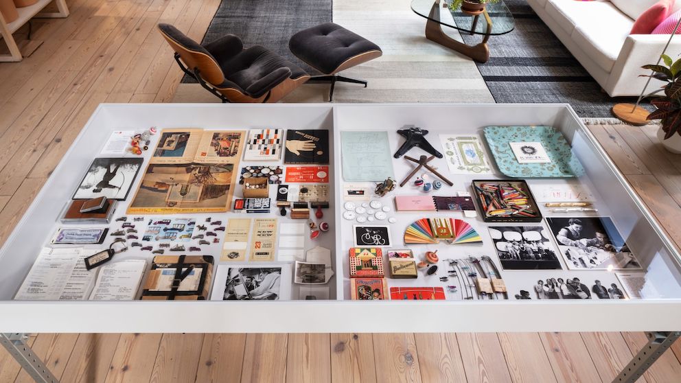 In the foreground, a display table holds two trays of archival objects like jacks, playing cards, crayons, and photographs. In the foreground, a display table holds two trays of archival objects like jacks, playing cards, crayons, and photographs.